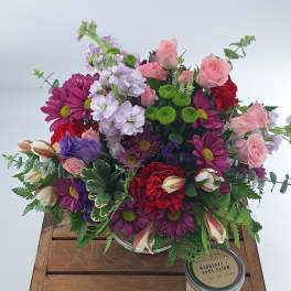 Mixed bouquet of pink roses, red carnations, purple daisies and green button mums in a glass vase with a candle beside it