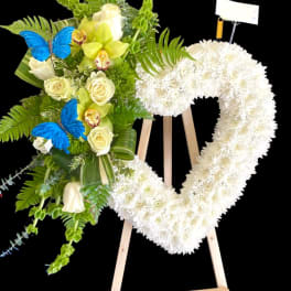 Heart-shaped white floral wreath on an easel beside a green and white bouquet with blue butterflies