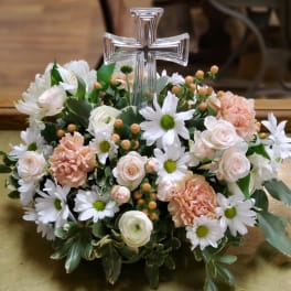 Floral arrangement with a clear cross centerpiece, white daisies, pale roses, and peach carnations