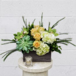 Arrangement of yellow roses, hydrangeas, orchids, and succulents in a rectangular container with a candle