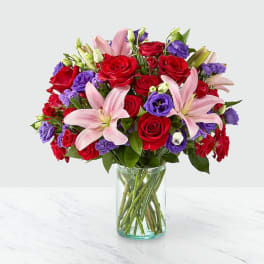 Bouquet of red roses, pink lilies, and purple blooms in a glass vase