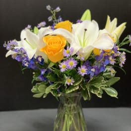 Bouquet of yellow roses, white lilies, and purple flowers in a glass vase