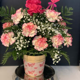 Pink and white carnations in a decorated pot with a heart pick