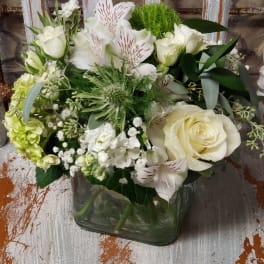 Compact white and green flower arrangement with roses in a clear glass cube vase on a rustic chair.
