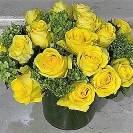Low arrangement of yellow roses with green accents in a clear glass cylinder vase