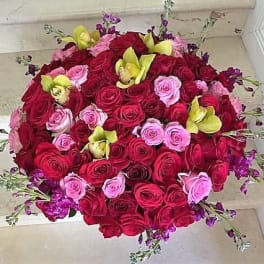 Dense round arrangement of red and pink roses accented with yellow orchids and small purple flowers