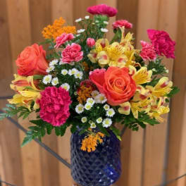 Bright mix of orange roses, pink carnations, yellow alstroemeria, and daisies in a blue glass vase.