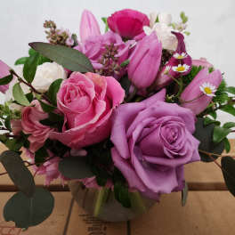 Low round arrangement of pink and lavender roses, tulips, and small white daisies in a clear glass bowl vase