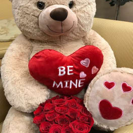Large teddy bear holding a red heart pillow and a box of red roses