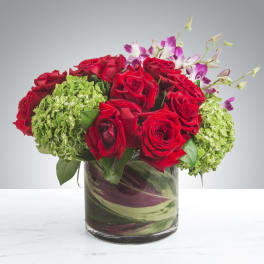 Low arrangement of red roses, green hydrangeas, and purple orchids in a leaf-wrapped glass cylinder vase