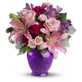Pink lilies, roses, and purple blooms arranged in a purple glass vase