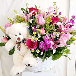 Pink and white flower arrangement in a white basket with a teddy bear