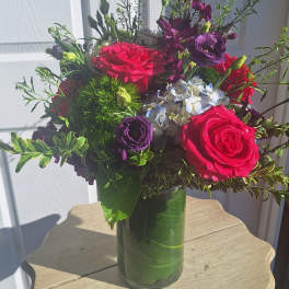 Vibrant arrangement of hot pink roses, purple flowers, and hydrangea in a clear glass vase.