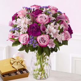 Pink and purple bouquet in a clear glass vase beside a box of chocolates