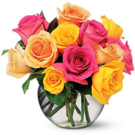 Short round arrangement of pink, yellow, and peach roses in a clear glass bowl vase