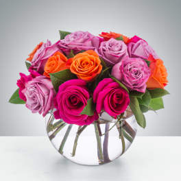 Compact arrangement of pink, lavender, and orange roses in a round glass vase