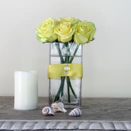Yellow roses in a clear rectangular vase with a yellow ribbon and candle beside it