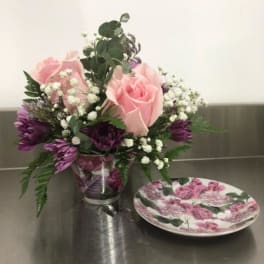 Pink roses and purple flowers in a clear vase beside a floral plate