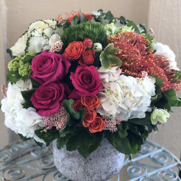 Low round arrangement of pink roses, orange spray roses, protea, and white hydrangeas in a gray pot