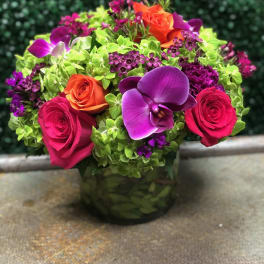 Round arrangement of bright roses, orchids, and green hydrangeas in a low glass vase