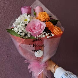 Hand-tied bouquet of pink roses, orange roses, white daisies and baby’s breath wrapped in pink paper.