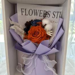 Boxed arrangement with orange rose, blue blooms, and cotton in lavender wrap with white ribbon
