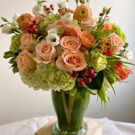 Tall vase arrangement of peach roses, green hydrangeas, white blooms and red berries.