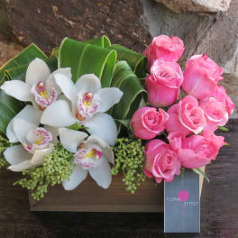 Pink roses and white orchids arranged with broad green leaves in a wooden box