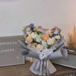 Pastel bouquet of roses, carnations, and blue flowers wrapped in gray paper