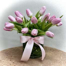 Arrangement of pink tulips in a clear glass vase wrapped with leaves and tied with a pink ribbon.