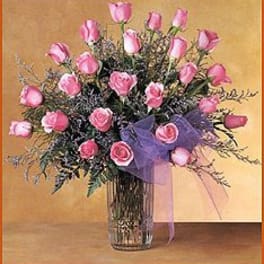 Tall arrangement of pink roses with purple accents in a clear glass vase with a purple bow