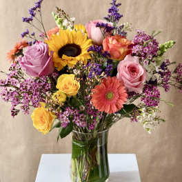 Mixed bouquet of roses, sunflower, and gerbera daisies in a glass vase