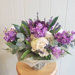 Hand-tied bouquet of purple stock and cream roses wrapped in pale paper on a wooden stool.