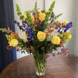 Mixed bouquet of yellow roses, blue flowers, and pink accents in a glass vase