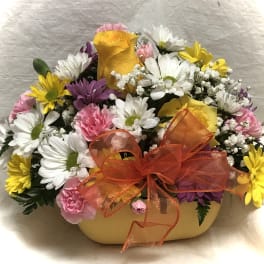 Mixed bouquet of daisies, roses, and carnations in a yellow container with an orange bow