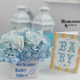 Blue and white flower arrangement in a white container with a baby card