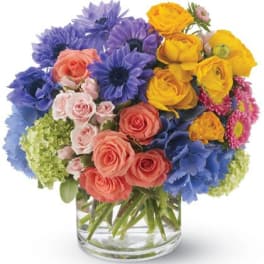 Bright mixed arrangement of blue, yellow, and peach flowers in a clear glass cylinder vase