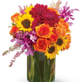 Bright arrangement of orange roses, yellow sunflowers, red blooms and purple orchids in a glass vase.