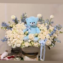 Blue teddy bear in a white rose and blue flower arrangement