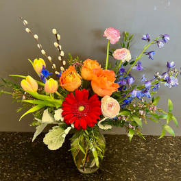 Mixed bouquet in a glass vase with red gerbera, orange roses, yellow tulips, and purple flowers