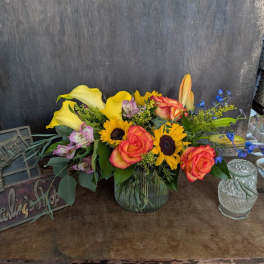 Mixed bouquet with roses, sunflowers, calla lilies, and blue accents in a glass vase