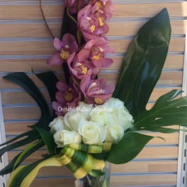 Pink orchids and white roses in a clear glass vase with large tropical leaves