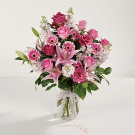 Pink roses, lilies, and gerbera daisies in a clear glass vase with a dotted ribbon