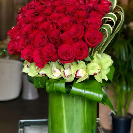 Tall arrangement of red roses with green cymbidium orchids in a leaf-lined green glass vase