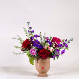 Asymmetrical arrangement of red roses and purple flowers in a tan ceramic face vase.