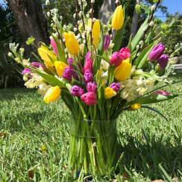 Tall glass vase of yellow and pink tulips with white flowers and pussy willow branches on grass