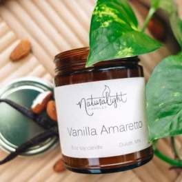 Brown glass Vanilla Amaretto candle jar with white label on a tray beside almonds, vanilla beans, and a green vine.
