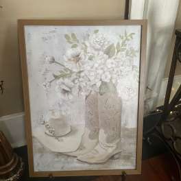 Framed artwork of white flowers in a vase beside a cowboy hat and boots