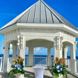 Two tropical floral arrangements flank a white cocktail table in a seaside pavilion
