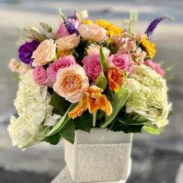 Mixed bouquet of pink, peach, purple, and yellow flowers in a square vase